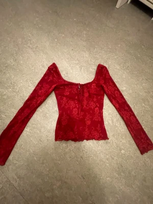Röd spetsblus från Gina Tricot - Snygg röd blus från Gina Tricot i spets med blommigt mönster. Modellen har lång ärm, croppad passform och en liten snörning framtill vid urringningen. Perfekt för dig som vill sticka ut lite extra.