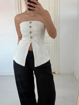 Vit linnetopp med knappar - Snygg vit axelbandslös linnetopp med knappar framtill och lätt utsvängd nederkant. Toppen har en bandeauform och är tillverkad i linne, vilket ger en fräsch och somrig känsla. Perfekt för dig som gillar stilrena och trendiga plagg.
