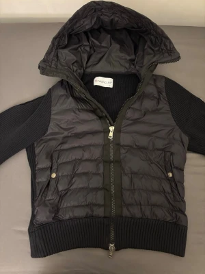 Svart dunväst med huva från Moncler - Snygg svart dunväst från Moncler med stickade svarta ärmar och stor huva. Västen har två fickor med tryckknappar, dragkedja framtill och Moncler-logga på ärmen. Materialmix av dun och stickat ger en cool look. Perfekt för lager på lager och streetstyle.