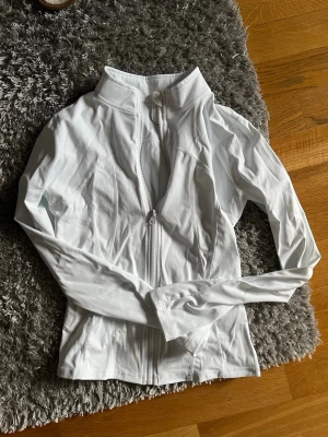 Vit sportjacka med dragkedja - Snygg vit sportjacka med hel dragkedja framtill och hög krage. Tillverkad i ett stretchigt och lätt material som passar perfekt till träning eller löpning. Figurnära passform och långa ärmar för en sportig look.