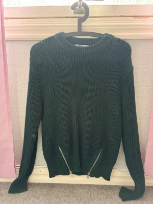 Mörkgrön stickad tröja med dragkedjor - Snygg mörkgrön stickad tröja med rund halsringning och två silverfärgade dragkedjor framtill. Tröjan har lång ärm och ribbstickade muddar vid hals, ärmslut och nederkant. Perfekt för dig som gillar en enkel men cool stil.