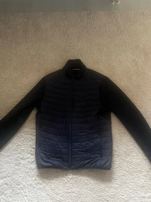 J.Lindeberg cardigan - Snygg svart pufferjacka med horisontella quiltade partier på fram- och baksida. Jackan har hel dragkedja, ståkrage och två sidofickor med dragkedja. Materialet är troligtvis syntet med lätt vaddering, perfekt för kalla vinterdagar. Den är som i nytt skick!