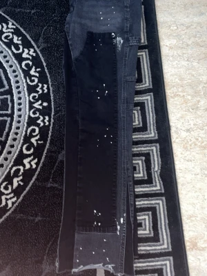 Svarta jeans med färgstänk - Unika svarta jeans med vita färgstänk och patchwork-detaljer längs benen. Raka ben och trendig look som sticker ut. Perfekta för dig som gillar streetwear och vill ha något som syns i mängden.