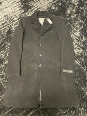 Svart ullkappa från Rhude ( Moose) storlek L - Stilren svart ullkappa från Rhude med klassisk krage i fårskinnsimitation och knappstängning framtill. Kappan har två snedställda fickor och detaljer med dragkedjor baktill samt en patch med texten 'Rhude Unchules' på ärmen.