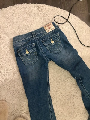 Blå True Religion jeans med kontrastsömmar - Säljer ett par klassiska blå jeans från True Religion med ikoniska kontrastsömmar och snygga fickdetaljer bak. Jeansen har låg midja, guldiga knappar. Perfekta för dig som gillar lågmidjade byxor.
