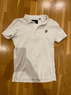 Vit pikéskjorta från Lyle & Scott - Vit pikéskjorta från Lyle & Scott med klassisk krage, korta ärmar och knappar framtill. Bröstet har den ikoniska gula fågel-loggan. Tillverkad i mjuk bomull för en bekväm och stilren look.