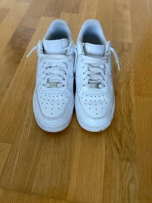 Vita Nike Air Force 1 sneakers - Klassiska vita Nike Air Force 1 sneakers i lågt utförande. Skorna har en stilren design med perforerad tå, vita skosnören och den ikoniska swoosh-loggan på sidan. Tillverkade i läder med gummisula för bra komfort och streetstyle-vibe.
