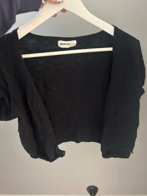 Svart kort kofta från Brownie - Kort svart kofta från Brownie med öppen front och lös passform. Tillverkad i ett tunt, mjukt material som ger en avslappnad look. Perfekt att slänga över en topp eller klänning för en enkel layering.