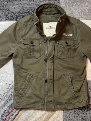 Olivgrön vindjacka från Hollister - Snygg olivgrön jacka från Hollister med klassisk krage, fyra fickor framtill och både knappar och dragkedja. Jackan har en stilren look och är perfekt för dig som gillar en avslappnad men trendig stil.
