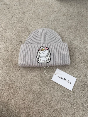 Acne Studios X Moomin Mössa i Ull - Säljer en ljusgrå ribbstickad mössa från Acne Studios med en stor patch framtill som föreställer en Muminfigur med hjärtan. Mössan har uppvikt kant och är tillverkad i mjukt material, perfekt för kalla dagar.