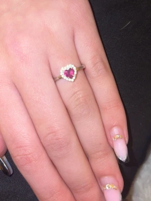 Silverring med hjärtformad röd sten - Söt silverfärgad ring med en hjärtformad röd sten i mitten, omgiven av små vita diamanter som ger en elegant touch. Perfekt för dig som gillar romantiska detaljer och vill ha något unikt på fingret.