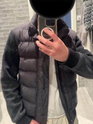 Svart Woolrich pufferjacka med stickade ärmar - Snygg svart pufferjacka med quiltad front och stickade ärmar. Jackan har hög krage, dragkedja och knappar framtill samt ribbade muddar vid ärmsluten. Perfekt för kalla vinter/höst/vår och ger en modern look. Jackan är köpt på woolrich i Paris och är i storlek small.