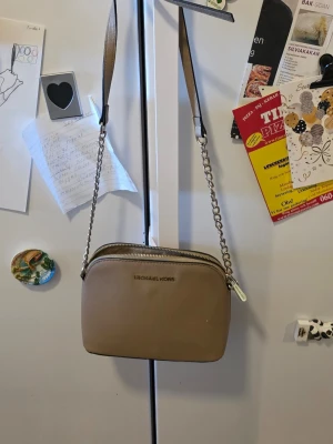 Beige handväska från Michael Kors - Snygg beige handväska från Michael Kors i ett tåligt, strukturerat läder. Väskan har guldfärgade detaljer, kedjerem och dragkedja med logotyp. Perfekt storlek för det viktigaste och stilren design som passar till många outfits.