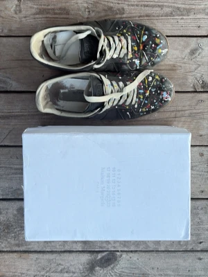 Maison Margiela sneakers med färgstänk - Unika Maison Margiela sneakers i svart läder med vita snören och färgstänk i flera färger över hela ovandelen. Klassisk låg modell med gummisula och ikonisk design. Perfekta för dig som vill sticka ut med din stil.