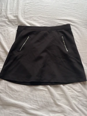 Svart kortkjol med dragkedjor från Lindex - Svart kortkjol från Lindex med två snygga dragkedjor framtill. Kjolen har en enkel och stilren design som passar till många olika outfits. Perfekt för dig som gillar en klassisk look med en liten edge.