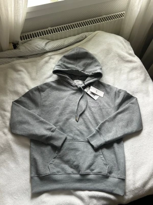Grå hoodie från Ami Paris - Snygg grå hoodie från Ami Paris med klassisk känguruficka framtill och justerbar dragsko i huvan. Tillverkad i mjuk bomullsblandning för maximal komfort. Perfekt för en avslappnad och stilren look. 