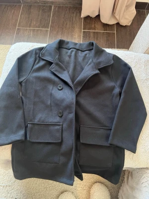 Svart klassisk kappa med fickor - Stilren svart kappa med två stora framfickor och två knappar framtill. Klassisk krage och rak passform ger en tidlös look. Perfekt för dig som vill ha en enkel men snygg jacka till hösten eller vintern. 