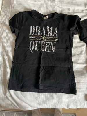 Svart t-shirt Drama Queen tryck - Svart t-shirt från Drama Queen med stort vitt och zebramönstrat tryck på bröstet där det står 'DRAMA QUEEN'. Klassisk rund hals och korta ärmar. Perfekt för dig som vill sticka ut med ett coolt statement-plagg.