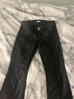 Svarta coated jeans från Nelly  - Säljer ett par svarta coated jeans från Nelly med låg midja och utsvängda. Byxorna har två silverknappar framtill och pressveck längs benen för en snygg look. Materialet har en lätt glansig finish som ger en cool vibe. Har inte använt