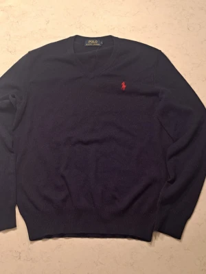 Mörkblå stickad tröja Polo Ralph Lauren - Mörkblå stickad tröja från Polo Ralph Lauren med klassisk röd logga broderad på bröstet. Tröjan har V-ringning, långa ärmar och ribbade muddar vid ärmslut och nederkant. Perfekt för dig som gillar stilren och tidlös design.