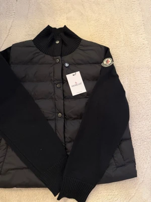 Svart dunväst från Moncler - Snygg svart dunväst från Moncler med stickade svarta ärmar och hög ribbad krage. Västen har tryckknappar framtill och Moncler-logga på ärmen. Perfekt för dig som vill ha en trendig och varm look i våren. Pris kan diskuteras vid snabbaffär 