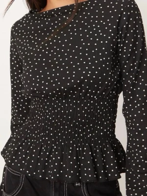 Svart prickig blus med smock - Trendig svart blus med vita små prickar och smockad midja som ger en snygg siluett. Blusen har långa ärmar och en lätt volang nedtill. Tillverkad i ett mjukt, vävt material som känns bekvämt mot huden. Perfekt att styla med både jeans och kjol.