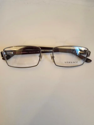 Versace glasögon med metallbåge - Snygga glasögon från Versace med rektangulära linser och metallbåge i silver. Skalmarna är sköldpaddsfärgade med Versace-logga i metall på sidan. Klassisk och stilren design som passar till många looks.