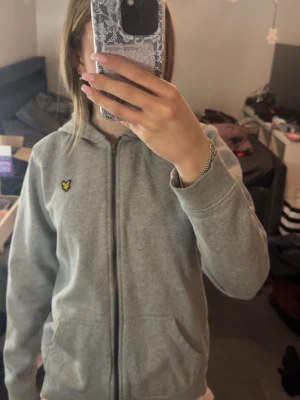 Grå zip hoodie från Lyle & Scott - Säljer en grå hoodie med dragkedja från Lyle & Scott. Klassisk modell med huva, två fickor framtill och den ikoniska gula örnloggan på bröstet. Perfekt till chill dagar och enkel att matcha med allt. Mjuk bomullskänsla och ribbade muddar.
