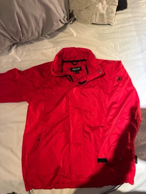 Röd vindjacka från Abacus - Snygg röd vindjacka från Abacus med hög krage och dragkedja framtill. Jackan har svarta detaljer, justerbar nederkant och diskret logga på bröstet. Tillverkad i lätt och tåligt syntetmaterial, perfekt för blåsiga dagar. Storlek 13-14 år 