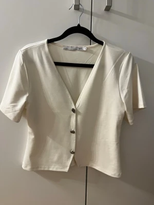 Vit v-ringad topp från Lager 157 - Stilren vit topp från Lager 157 med v-ringning och korta ärmar. Toppen har tre knappar framtill och är lätt figursydd för en snygg passform. Perfekt att matcha med jeans eller kjol för en enkel och fräsch look.