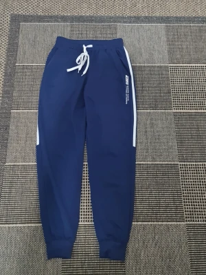 Blå mjukisbyxor med vita detaljer - Snygga blå mjukisbyxor från RED med vita sidoränder och dragsko i midjan. Tryck på benet med texten 'BURNING PASSION SURPASS DREAMS'. Perfekta för chill dagar eller träning. Mjuk resår i benslut för skön passform.