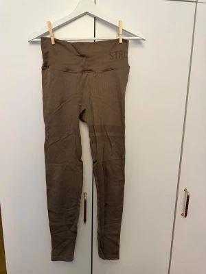 Ribbade bruna leggings STRONGER - Säljer ett par ribbade leggings från STRONGER i en snygg brun färg. De har hög midja och är tillverkade i ett stretchigt, mjukt material som sitter skönt på kroppen. Perfekta för träning eller chill. Snyggt diskret mönster och logga i midjan.