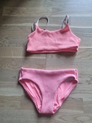Rosa bikini från H&M - Säljer en söt rosa bikini från H&M i storlek 146/152. Bikinin har en ribbad struktur och består av ett sportigt linne med smala axelband och matchande trosor. Perfekt för lata dagar på stranden eller poolen!