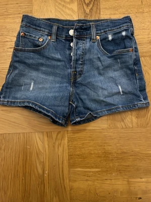 Blå jeansshorts från Levi's - Säljer ett par klassiska blå jeansshorts från Levi's med hög midja och slitna detaljer. Shortsen har fem fickor, knappgylf. 