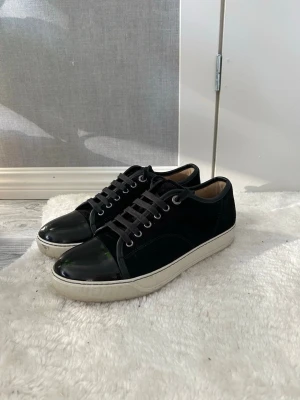 Svarta sneakers från Lanvin - Snygga svarta sneakers från Lanvin med ovandel i mocka och detaljer i blankt läder på tån och hälen. Skorna har vita gummisulor och svarta skosnören. Klassisk låg modell som passar till det mesta och ger en clean look.