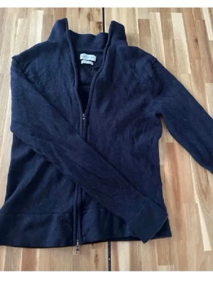 Svart ullkofta med dragkedja - Kofta i svart ull med hel dragkedja framtill och hög krage. Klassisk och enkel design som passar till det mesta. Långa ärmar och mjukt material för extra komfort.