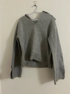 Grå stickad hoodie med v-ringning - Mysig grå stickad tröja med luva och v-ringad hals. Tröjan har långa ärmar, bred ribbstickad mudd vid ärmslut och nederkant samt en avslappnad passform. Perfekt för lager på lager och chill dagar.