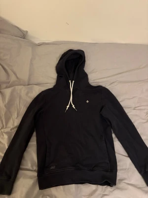 Svart hoodie från Morris - Snygg svart hoodie från Morris med vit dragsko i huvan och diskret logga på bröstet. Tillverkad i mjukt bomullsmaterial och har ribbade muddar vid ärmslut och nederkant. Perfekt för en avslappnad och stilren look.