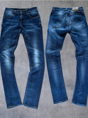 Nudie Jeans 29/32 - Snygga blåa jeans från nudie med slitna detaljer och tvättade partier på låren och knäna. Klassisk femficksmodell med knappgylf och raka ben. Perfekta för en avslappnad och trendig look. Bara och ställa frågor😉