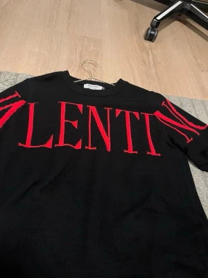 Svart stickad tröja från Valentino - Snygg svart stickad tröja från Valentino med stora röda bokstäver över bröstet och detaljer på ärmarna. Rund halsringning och ribbade muddar. Tillverkad i Italien, perfekt för dig som gillar exklusiva och trendiga plagg.
