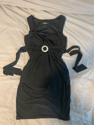 Svart kort klänning från BCX - Snygg svart kort klänning från BCX med breda axelband och dekorativt spänne i midjan. Klänningen har omlott-detalj upptill och knytband i midjan för en markerad siluett. Perfekt för dig som vill ha en elegant och stilren look.