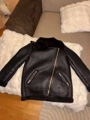 Svart skinnjacka med sned dragkedja - Säljer en svart skinnjacka med cool sned dragkedja och två fickor med dragkedjor framtill. Jackan har en hög krage med spänne och en stilren, lite boxig passform. Perfekt för dig som vill ha en edgy look till hösten eller vintern.