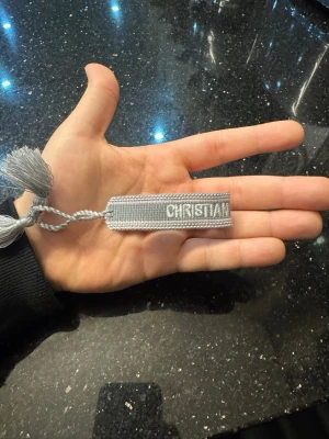 Grått Christian Dior J'Adior armband - Snyggt grått armband från Christian Dior med texten 'Christian Dior J'Adior' broderat i vitt. Armbandet har justerbar knytning och dekorativa tofsar i ändarna. Perfekt accessoar för dig som gillar exklusiv och trendig stil.