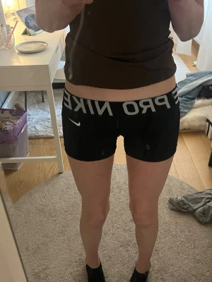 Svarta Nike Pro shorts - Säljer ett par svarta Nike Pro shorts med bred resår i midjan och vit Nike-logga på sidan. Shortsen är tighta och har en sportig look, perfekta för träning eller gym. Materialet är stretchigt och bekvämt. Det står att det är storlek m men jag skulle säga xs/s men funkar så klart med m också eftersom de är stretchiga! Hör av vid frågor💗💗