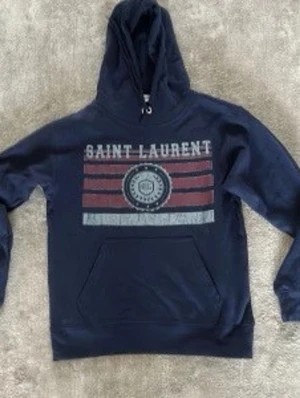 Mörkblå Saint Laurent hoodie - Snygg mörkblå hoodie från Saint Laurent med tryck framtill i vitt och rött. Klassisk känguruficka och justerbar huva med snören. Perfekt för en chill och avslappnad stil. Materialet känns mjukt och bekvämt.