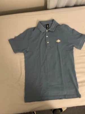 Ljusblå pikétröja från replay - Snygg ljusblå pikétröja från J.Lindeberg med klassisk krage och tre knappar framtill. Tröjan har korta ärmar och en liten vit logga på bröstet. Perfekt för en clean och stilren look.