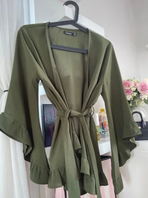 Olivgrön omlottsblus från Boohoo - Snygg olivgrön omlottsblus från Boohoo med volangkant och vida ärmar. Knytskärp i midjan för en avslappnad look. Tillverkad i ett mjukt, lätt material som känns skönt mot huden. 