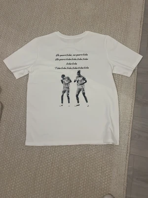 Vit t-shirt med tryck och text - Vit t-shirt med svart tryck på ryggen som visar två personer och text ovanför. Klassisk passform och korta ärmar, perfekt för dig som gillar unika motiv på kläder. Snygg och enkel att matcha med jeans eller shorts. Helt ny, aldrig andvänd