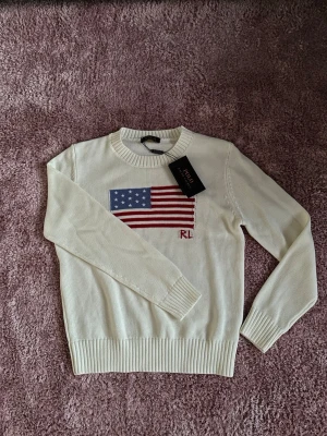 Vit stickad tröja Polo Ralph Lauren - Vit stickad tröja från Polo Ralph Lauren med amerikansk flagga broderad på bröstet och RL-detalj i rött. Tröjan har rund halsringning, långa ärmar och ribbade muddar vid ärmslut och nederkant. Klassisk och stilren design.