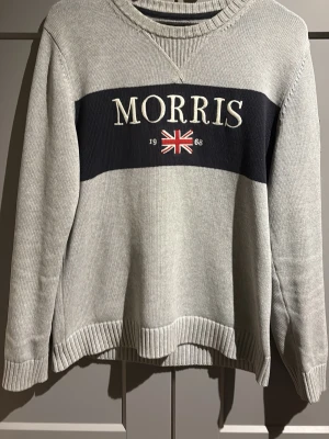 Grå tröja från Morris - Snygg grå tröja från Morris med stor logga och brittisk flagga broderad på bröstet. Tröjan har rund halsringning, ribbade muddar och en mörkblå panel framtill. Perfekt för en stilren och avslappnad look. Passar Medium   Priset kan förhandlas